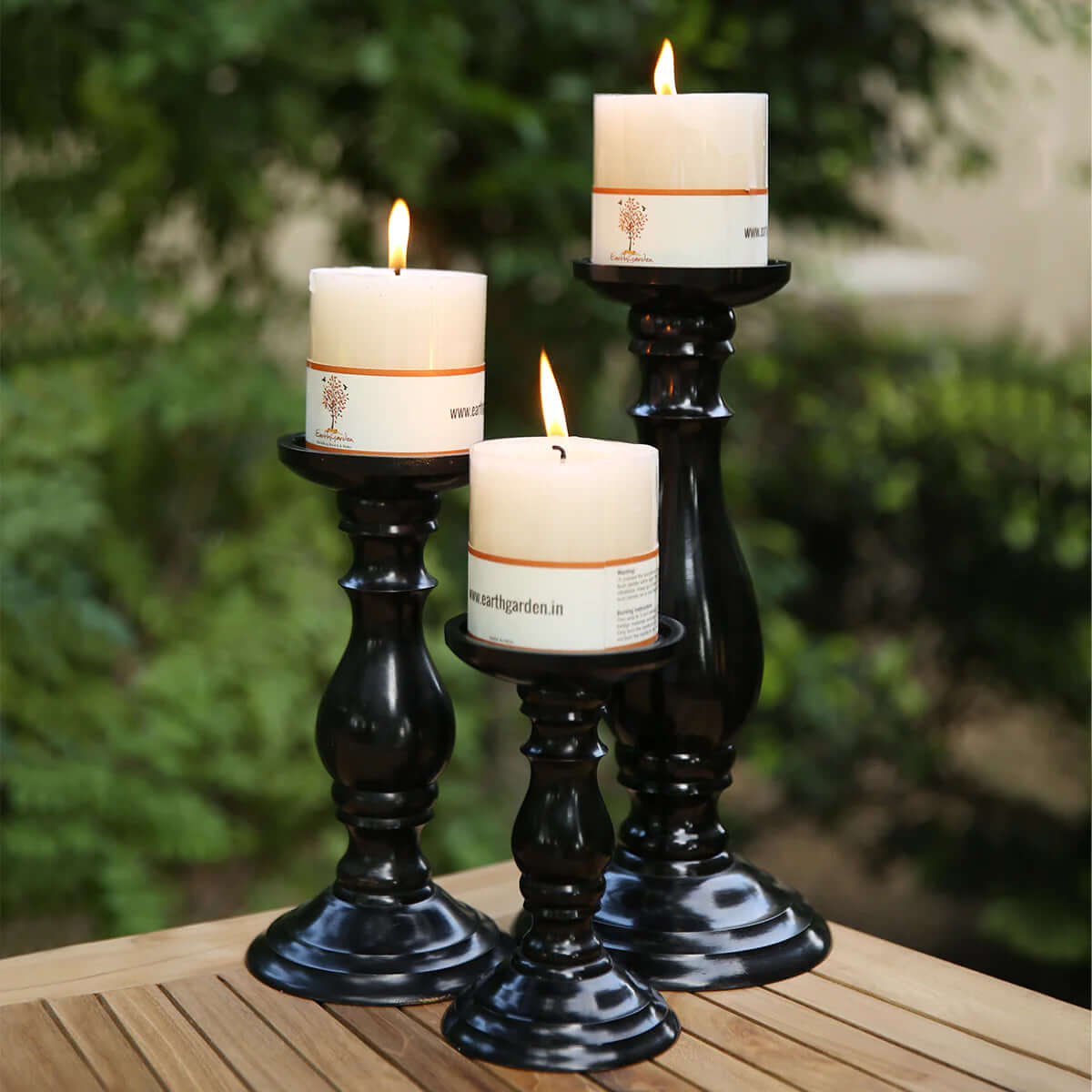 Three white pillar candles are lit and placed on traditional MSS Store 01 Candle Holder 2 of varying heights. They are arranged on a wooden table, with a blurred leafy background, creating a warm and elegant home decor ambiance.