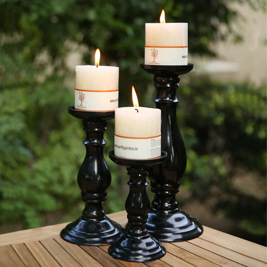 Three white pillar candles are lit and placed on traditional MSS Store 01 Candle Holder 2 of varying heights. They are arranged on a wooden table, with a blurred leafy background, creating a warm and elegant home decor ambiance.